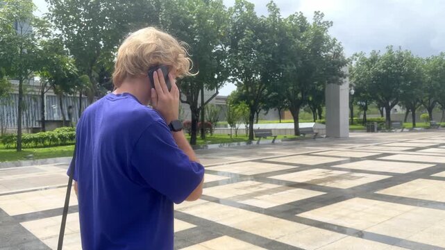 Young Blond Man Walking on Wide Park Path in Shenzhen and Talking on Smartphone &ndash; Back View with Tropical Greenery and Blue Sky