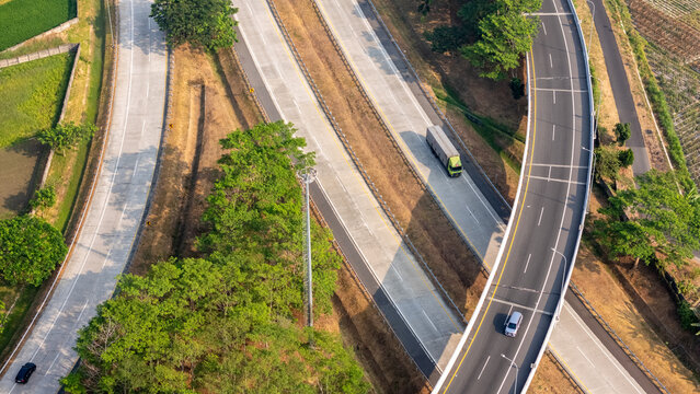 Aerial view of a highway interchange with a truck and a car driving on the road surrounded by lush green trees and agricultural fields in Boyolali, Central Java, Indonesia.