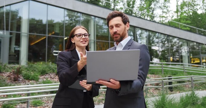 Businessman and businesswoman smiling and discussing work while looking at a laptop screen outdoors. Teamwork, business communication, collaboration, and modern work lifestyle.