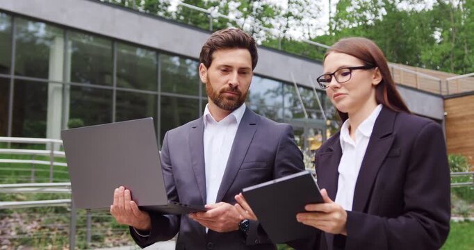 Businessman and businesswoman walk to the office while having a business conversation and looking at a tablet held by the woman, with the man carrying a laptop.