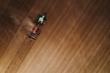 High top-down aerial angle of a green tractor and seeder driving diagonally across a vast brown field, generating a subtle trailing cloud of dust. © eddgars