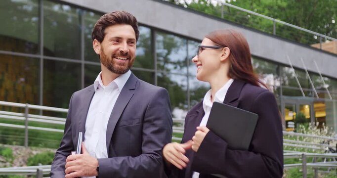 Businessman and businesswoman walking through a city park, smiling and talking on their way to the office.