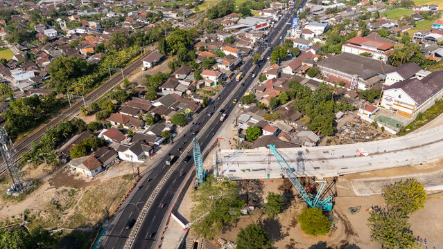 Aerial view of a bridge construction site with cranes and concrete structures near a busy road, railway tracks, and houses in Yogyakarta, Special Region of Yog, Indonesia.