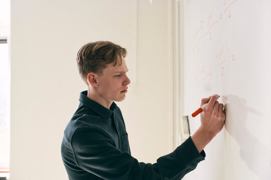 Focused teenage male student solving mathematics equation on whiteboard in classroom at college