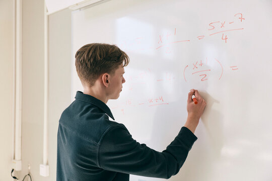 Focused teenage male student solving mathematics equation on whiteboard in classroom at high school
