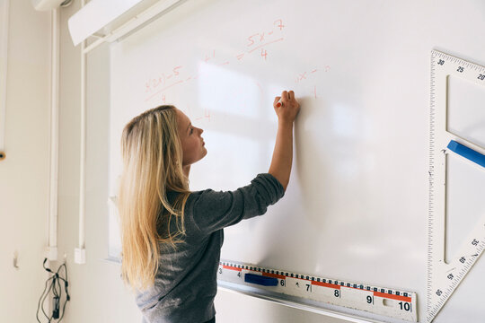 Blond teenage girl solving mathematics equation on whiteboard in classroom at college
