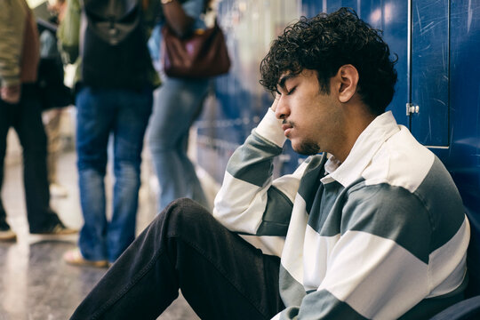 Depressed male college student sitting with eyes closed in corridor near lockers