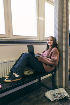 Portrait of smiling teenage female student sitting with laptop on bench in college corridor