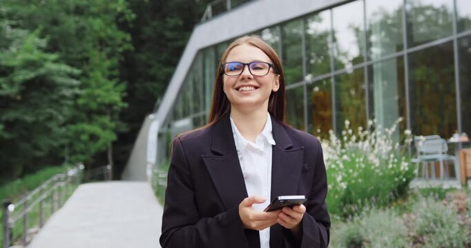 Young woman walking through a city park, smiling and using her smartphone to exchange text messages with friends.