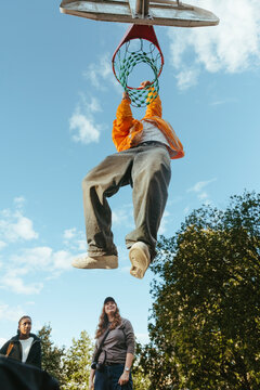 Low angle view teenage boy hanging from basketball rim after scoring dunk in school playground area