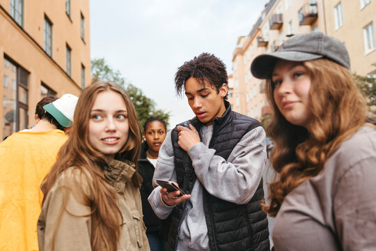 Teenage boy using smart phone while standing amidst friends in school campus