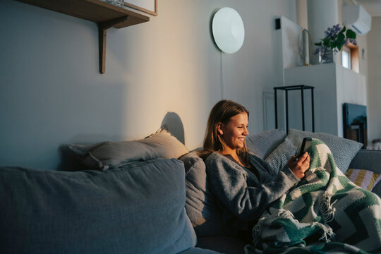 Smiling teenage girl spending leisure time using smart phone while sitting on sofa at home