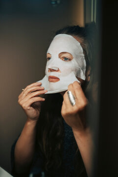Mature woman peeling off skincare mask while looking in mirror at home