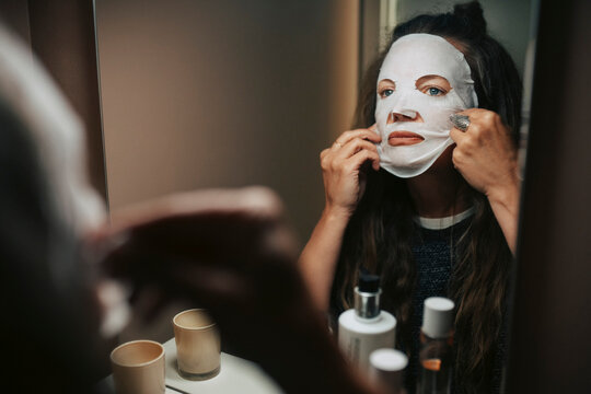 Mature woman applying skincare face mask while looking in mirror