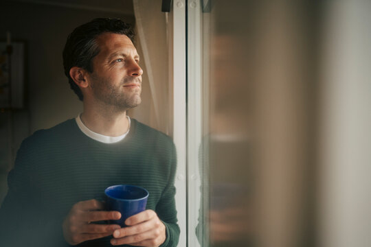 Thoughtful man holding blue mug while looking out of window