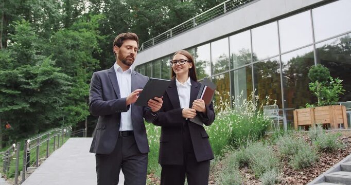 Young businesswoman and businessman walking through a city park, discussing work while looking at a tablet screen held by the man.