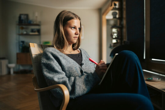 Thoughtful girl sitting with pen and book on chair at home