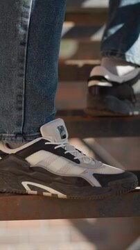 Close up of person feet wearing white and black sneakers on metal stairs. shoes are detailed with mesh fabric laces branding patches rubber soles denim jeans sunlight casting shadows