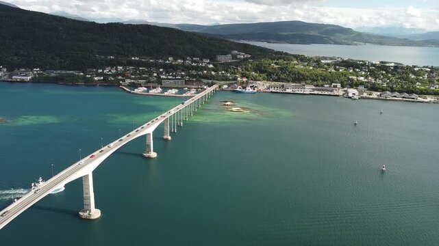Aerial drone footage captures a fishing boat passing beneath the Gisund Bridge in Senja, Norway, while cars cross above and the bridge connects the island to the mainland town of Finnsnes