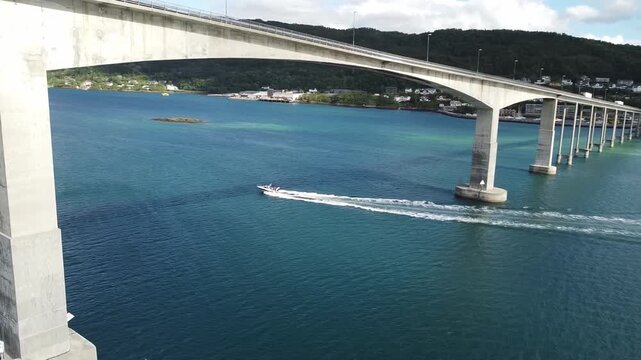 A speedboat passes beneath the Gisund Bridge in Senja, Norway, leaving a long wake trail across the calm blue water below the towering concrete piers, Drone orbit shot