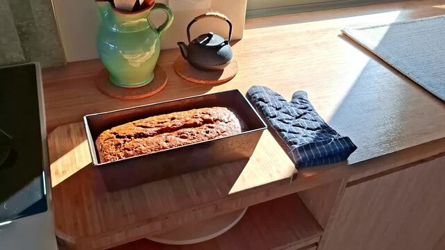 A freshly home baked banana bread in a baking tin on a breadboard in a kitchen with an oven glove as warm light filters through the window
