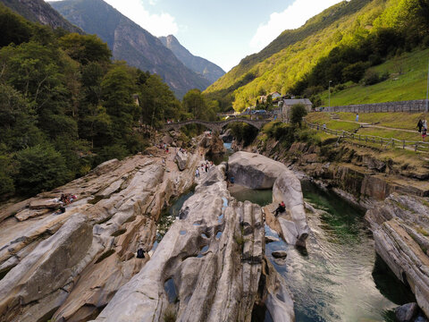 Aerial view of Lavertezzo and the iconic stone bridge over the emerald Verzasca River, surrounded by lush mountains and smooth rock formations in Verzasca, Ticino, Switzerland.
