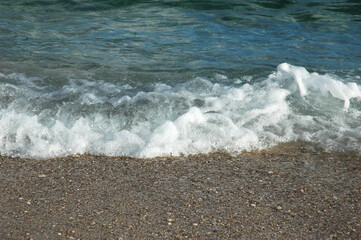 A close up shot of a turquoise sea wave with white splash foam hitting a wet pebble beach shore. © nahhan