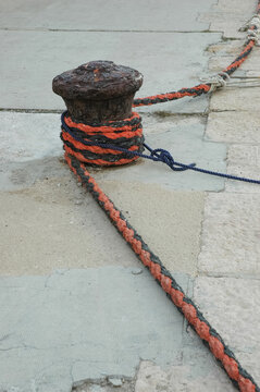 An old rusty iron mooring bollard is wrapped with heavy orange and black ropes on a traditional stone pier.
