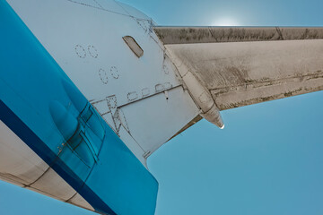 Low angle detail of a passenger jet tail fin and stabilizer against the sun, showing weathered panels and a bold blue livery stripe. © Victor