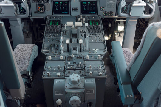 Interior of a classic airliner flight deck with throttle quadrant, radio panels with green displays and two pilot seats with sheepskin covers.