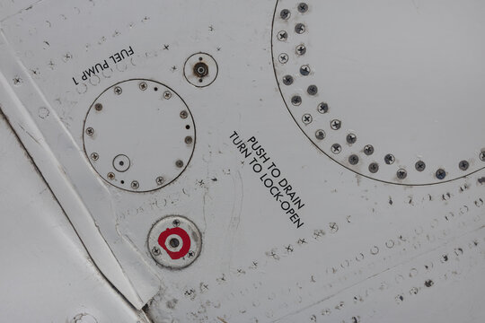 Abstract detail of white aircraft skin with fuel pump and drain valve markings, service panels, screws and rivets on a parked passenger jet.