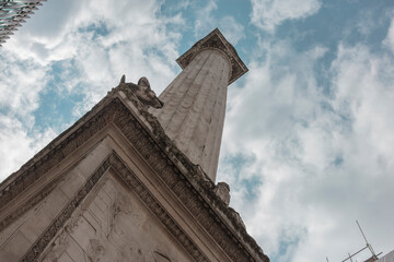Looking up the fluted Doric column of the Monument to the Great Fire of London, with a carved cornice and dragon against a cloudy sky. © Victor