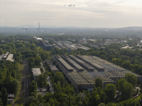 Aerial view of an industrial complex featuring large factory buildings, warehouses, and technical installations surrounded by lush green trees and railway tracks in Poland.