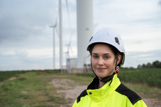 A confident female engineer wearing a hard hat and high visibility jacket stands outdoors at a wind farm with large turbine towers in the background, promoting green energy and sustainability.