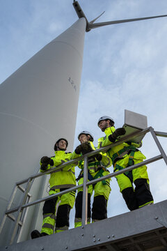 Team renewable energy engineers wearing high visibility jackets and safety helmets inspect a large wind turbine outdoors during a daytime maintenance project, focusing on modern green power.
