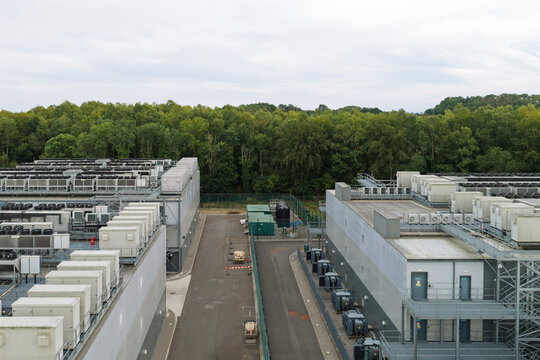 Aerial view of a data center facility featuring industrial cooling units on rooftops, a paved access road, and a dense forest background in United Kingdom.