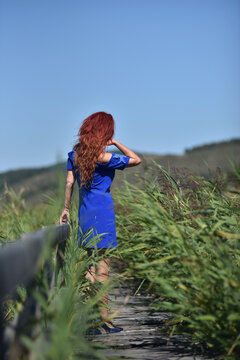 Red-haired woman in a vibrant blue dress, standing on a wooden footbridge
