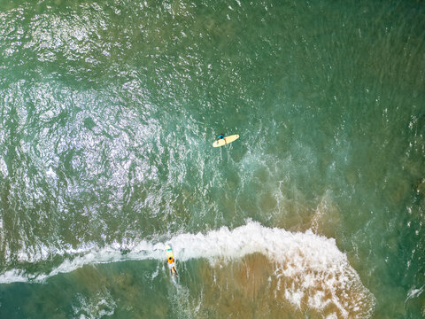 Aerial view of two surfers on surfboards in the clear turquoise waters of the Mediterranean Sea, with one surfer catching a wave and another paddling in Chania, Greece.