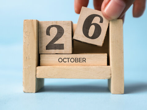 A woman's hand is holding a wooden block with the number 6 written on it on a wooden block calendar for October 26th on a light blue background.