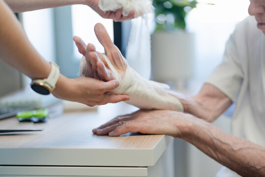 Caring for elderly patients with professional dedication, close-up bandaging both senior hands with gauze by compassionate nurse providing quality medical assistance in clinical setting