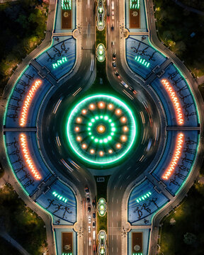 Aerial view of the illuminated Unirii Square Fountains at night, featuring green and orange water displays, light trails from traffic, and symmetrical landscaping in Bucharest, Romania.