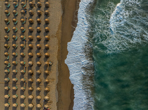 Aerial view of rows of straw umbrellas and sun loungers on the sandy beach with turquoise waves washing the shore in Chania, Greece.