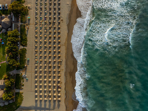 Aerial view of rows of straw umbrellas on the sandy beach with turquoise waves washing the shore in Chania, Greece.