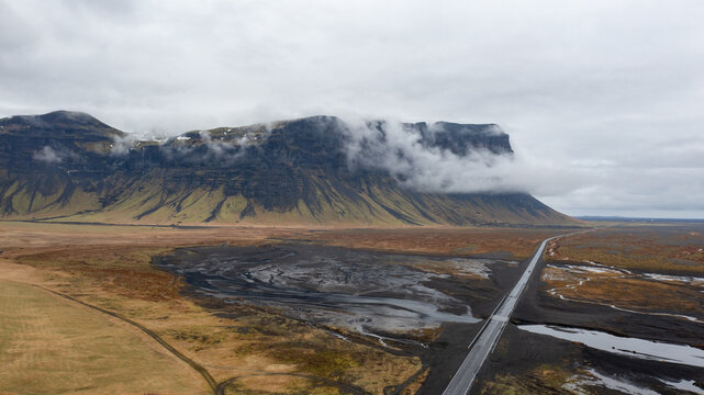 Aerial view of a long road stretching across a vast, dark volcanic plain beneath a massive mountain ridge partially covered by low-hanging clouds in Iceland.