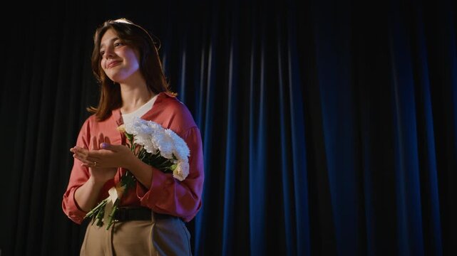Caucasian teenage girl holding flower bouquet and clapping hands on theater stage, low angle view. Perfect for drama school advertising and youth arts promotion