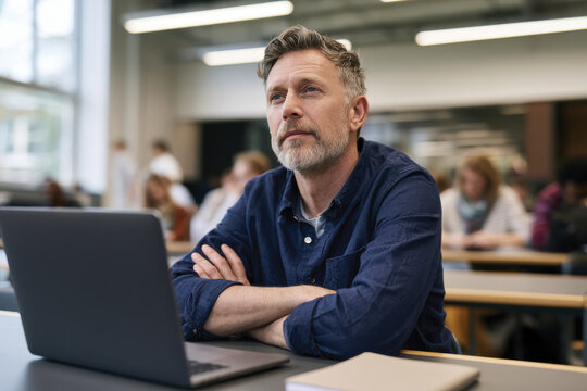 A thoughtful man sits in a classroom with a laptop, surrounded by students engaged in learning, reflecting concentration and a desire to acquire knowledge in a modern setting.