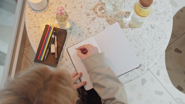 Woman with long blonde hair writes in notebook on round marble table selecting colored pencils from leather case soft lighting creates warm ambiance with water bottle and small flower vase