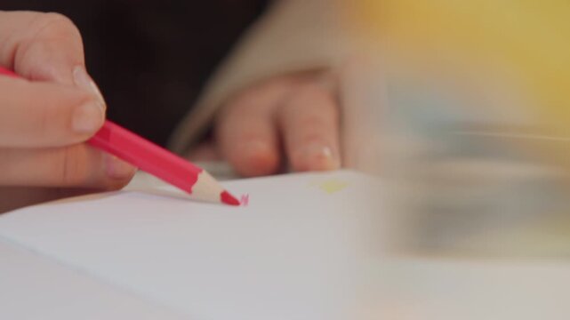 Close up of hand holding and using pink colored pencil to draw on white paper. person hands are visible with focus remaining sharp throughout as they create marks. Soft lighting highlights