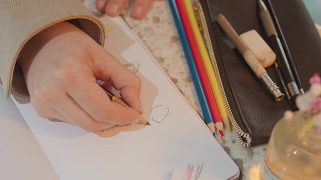 Close up of hand drawing on paper with colored pencils nearby. woman sketches simple shapes while various stationery items like pens and eraser rest beside notebook. Soft lighting highlights