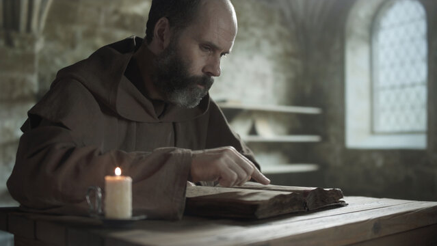 Medieval monk following lines with a finger while reading an open book at a wooden table near a burning candle inside a scriptorium